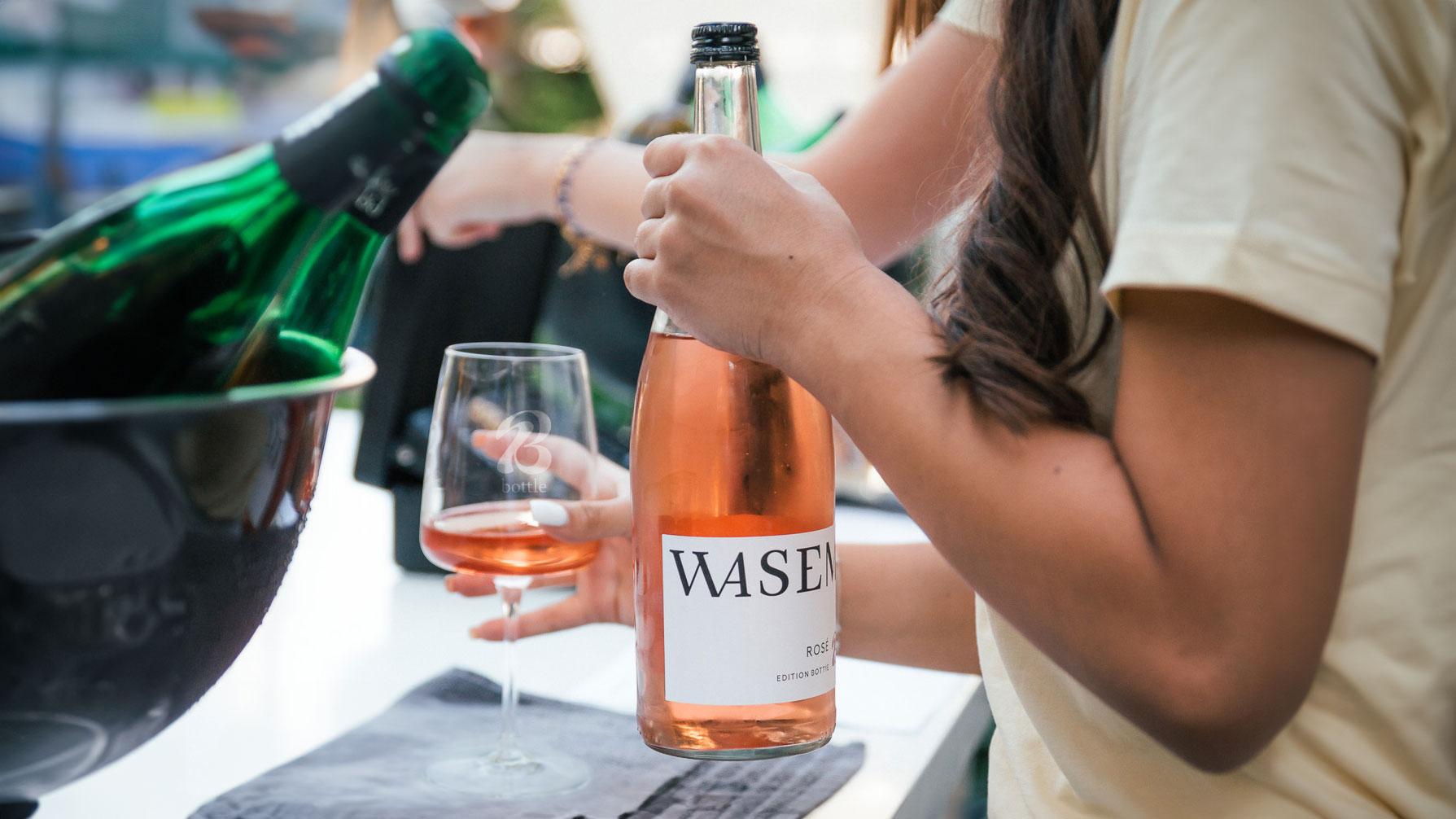 A woman holds a Bott1e glass filled with Wasem Rosé. In her other hand she holds the Wasem Rosé bottle.